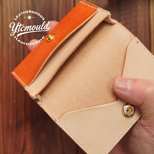 Puede incluir: Tarjetero de cuero abierto con solapa naranja e interior beige natural. El tarjetero presenta un cierre de botón a presión dorado y costuras blancas. El logotipo "LEATHERGOODS Ytcmould HANDMADE" es visible.