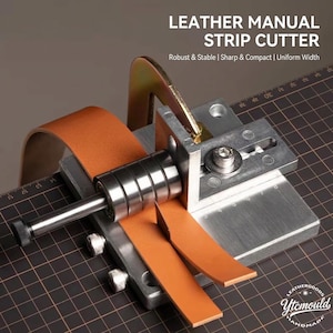 Op de afbeelding: Een handmatige leren stripsnijder met de tekst "LEATHER MANUAL STRIP CUTTER". De tool is gemaakt van metaal en heeft een mes, rollen en een geleider voor het snijden van leren strips. Een stuk bruin leer wordt gesneden.