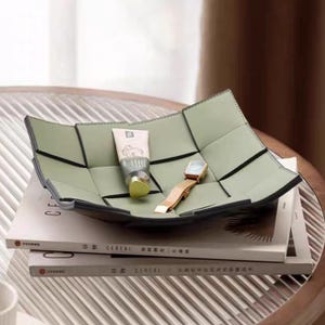 Puede incluir: Una bandeja verde salvia de estilo tejido con ribete negro, que contiene un tubo de crema y un reloj con correa dorada. La bandeja se encuentra sobre dos libros, con la palabra "CEREAL" visible en los lomos, sobre una mesa con tapa de cristal.