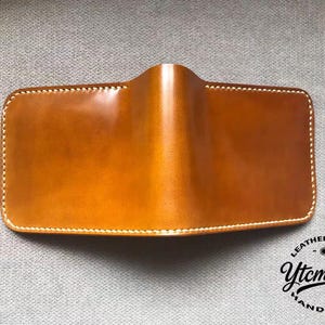 Könnte beinhalten: Rechteckige Ledergeldbörse in warmem Goldbraun. Sie hat weiße Nähte an den Kanten und ein geschwungenes Design oben. Das Logo "Ytcmould Handmade Leathergoods" ist unten rechts sichtbar.