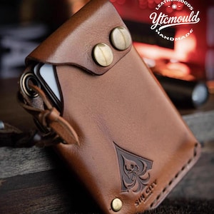 May include: A brown leather wallet with a flap secured by two brass-coloured snaps. The wallet features a decorative spade design with the text "SIN CITY" embossed on the front. A leather strap and a metal ring are attached to the side.