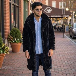 May include: A long, black faux fur coat with a wide collar. The coat is worn over a light blue button-down shirt and dark blue jeans. The person is wearing sunglasses and standing on a brick-paved sidewalk.
