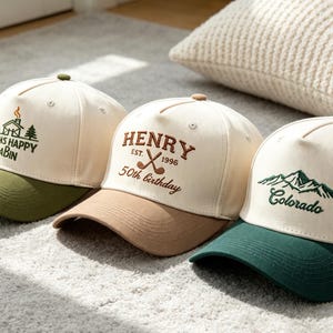 Puede incluir: Tres gorras de béisbol con diseños bordados. Una gorra tiene el texto "LUCAS HAPPY CABIN", otra dice "HENRY EST. 1996 50th Birthday", y la tercera "Colorado". Cada gorra tiene una corona color crema y una visera de color.