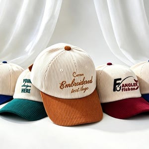 Pode incluir: Uma coleção de cinco bonés de beisebol de veludo cotelê com copas na cor creme e abas contrastantes em azul, verde, marrom e bordô. Cada boné apresenta textos e logotipos bordados exclusivos, incluindo designs de clubes de golfe.