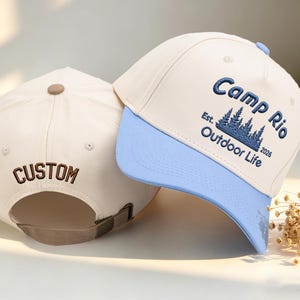 Peut inclure: Deux casquettes de baseball. L'une est de couleur crème avec "CUSTOM" brodé au dos. L'autre a une visière bleu clair et le texte "Camp Rio Outdoor Life" avec un motif d'arbre. Les deux ont des sangles réglables.
