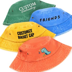 Könnte beinhalten: Vier Baumwoll-Fischerhüte in Grün, Blau, Gelb und Orange. Der grüne Hut zeigt den weißen Schriftzug CUSTOM Embroidery. Der blaue Hut trägt den schwarzen Schriftzug F.R.I.E.N.D.S. Der gelbe Hut zeigt CUSTOMIZE BUCKET CAP. Der orange Hut hat ein Dinosaurier.