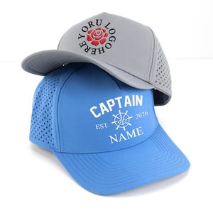 May include: Two baseball caps, one gray and one blue, are stacked. The gray cap has a rose logo and the text "YORU LOGO HERE". The blue cap features the text "CAPTAIN EST. 2026 NAME" with a nautical wheel graphic.