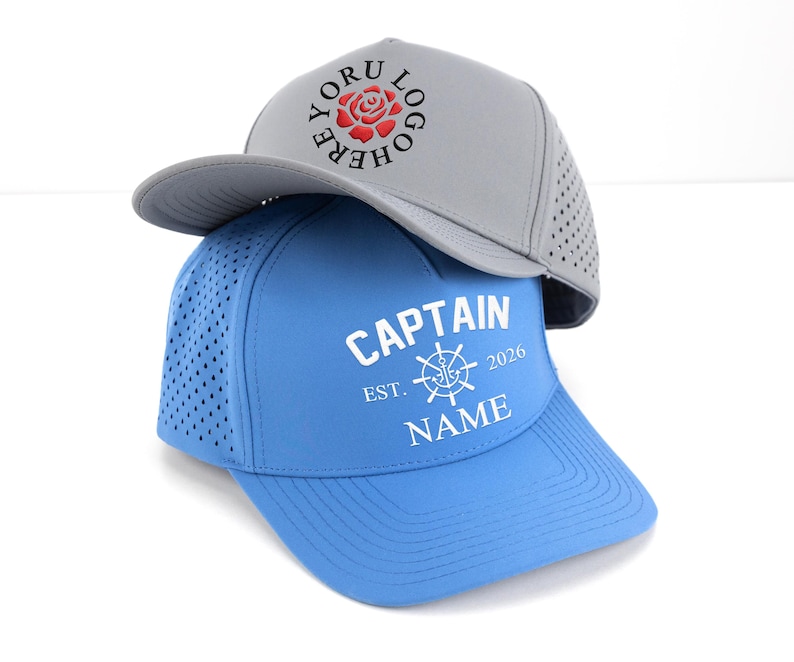May include: Two baseball caps, one gray and one blue, are stacked. The gray cap has a rose logo and the text "YORU LOGO HERE". The blue cap features the text "CAPTAIN EST. 2026 NAME" with a nautical wheel graphic.