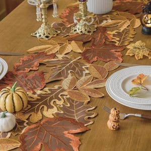 Puede incluir: Una mesa con temática otoñal con un camino de mesa en forma de hoja marrón con detalles bordados. La mesa está puesta con platos blancos, un tenedor y calabazas decorativas. Candelabros dorados y una pequeña linterna completan el ambiente otoñal.