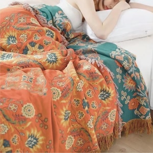 Könnte beinhalten: Eine orangefarbene und türkisfarbene Decke mit floralem Muster und Fransen. Die Decke zeigt ein detailliertes Design von Blumen und Blättern in Orange-, Türkis-, Gelb- und Blautönen. Die Decke ist über ein Bett drapiert.