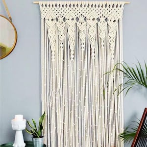 Könnte beinhalten: Cremefarbener Makramee-Wandbehang mit geometrischem Design. Die Wanddekoration zeigt eine Reihe von Rautenformen und vertikalen Strängen mit kleinen Knoten. Das Stück ist an einem Holzstab aufgehängt und verleiht jedem Raum einen Hauch von Bohème-Stil.