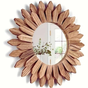 Op de afbeelding: Een ronde decoratieve spiegel met een sunburst-ontwerp. Het frame is gemaakt van lichtbruine houten stukken die zijn gerangschikt om op zonnestralen te lijken. De spiegel weerspiegelt een kamer met een raam en groen.