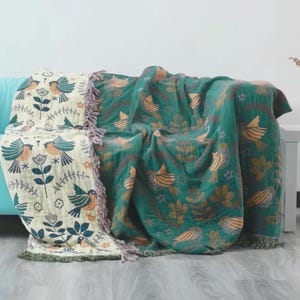 May include: Two decorative throw blankets with bird and floral patterns. One is cream with teal, orange, and navy blue birds and flowers. The other is emerald green with orange birds and brown leaves. Both have fringed edges.