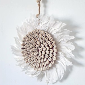 May include: A decorative wall hanging with a central circular design made of seashells. The seashells are surrounded by white feathers, creating a flower-like appearance. A natural fibre cord is attached for hanging.