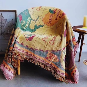 Può includere: Una coperta tessuta con un disegno di mappa del mondo drappeggiata su una sedia. La coperta ha uno sfondo giallo con continenti verdi, rossi e marroni ed è rifinita con un bordo a frange. Le parole "North America", "South America" e "Pacific Ocean" sono visibili.