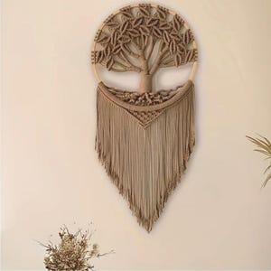 May include: A tan macrame wall hanging depicting a tree within a circular wooden frame. The tree design features a trunk, branches, and leaves, all made from knotted rope. The bottom of the piece has long, flowing fringe.