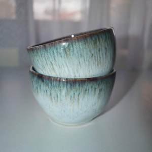 Puede incluir: Dos cuencos de cerámica apilados. Tienen un esmalte azul claro con detalles en azul oscuro y marrón en el borde. Los cuencos son probablemente para servir alimentos. Cada cuenco mide aproximadamente 12 cm de diámetro.