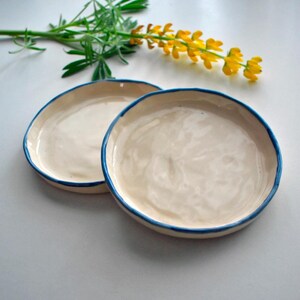 Può includere: Due piatti in ceramica bianco sporco con un bordo blu brillante. I piatti sono rotondi e hanno una forma leggermente irregolare. Un rametto di fiori gialli e foglie verdi sullo sfondo.