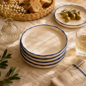 Può includere: Una pila di quattro piatti rotondi in ceramica con una superficie liscia color crema e un bordo blu scuro dipinto a mano. I piatti presentano una forma del bordo leggermente irregolare e organica, che mette in risalto il contrasto tra la base chiara e il bordo blu.