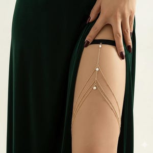 Può includere: Catena per gamba color oro con tre fili e dettagli di perle finte. Il gioiello per il corpo è indossato sulla coscia, con una fascia nera in alto. La catena è drappeggiata su una gamba, con un vestito verde scuro sullo sfondo.