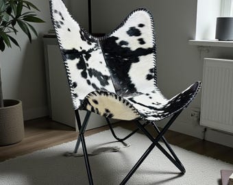 Könnte beinhalten: Ein Schmetterlingsstuhl mit einem schwarz-weißen Kuhfellbezug mit sichtbaren genähten Kanten, getragen von einem schwarzen Metallrahmen. Die Sitzfläche hat eine ausgeprägte Flügelform und besteht aus gemustertem Tierfellmaterial.
