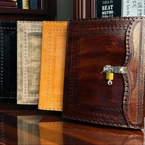 Puede incluir: Una colección de diarios encuadernados en cuero de varios colores, incluyendo negro, beige, amarillo y marrón oscuro. Los diarios tienen diseños en relieve intrincados y un cierre de cerradura. Los diarios se exhiben sobre una superficie de madera.