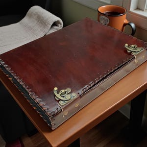 Puede incluir: Una caja rectangular de cuero marrón oscuro con cierres y costuras de color latón. La caja está sobre una pequeña mesa marrón, con una manta beige enrollada y una taza de té cerca. La caja parece ser un diario o un estuche de almacenamiento.