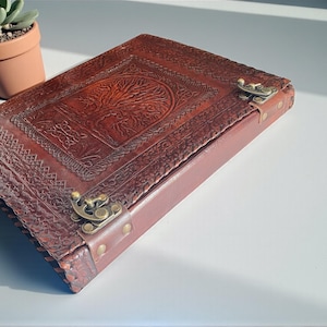Op de afbeelding: Een bruin leren notitieboek met een boom des levens-ontwerp in reliëf op de omslag. Het notitieboek heeft een sluiting met een sluiting en is afgewerkt met messing hardware. Op de achtergrond staat een kleine potplant.
