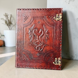 May include: A rectangular brown leather journal featuring an embossed design of two intertwined dragons with wings on the front cover. The cover is decorated with intricate border patterns and secured by two antique brass-colored metal latches.