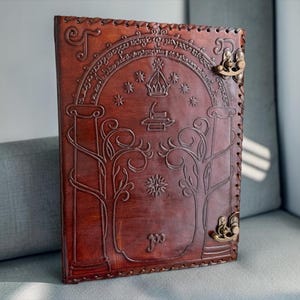 Könnte beinhalten: Ein rechteckiges Notizbuch aus braunem Leder mit einer Prägung von zwei Bäumen, einer Krone, Sternen und einem Torbogen mit Schriftzug. Der Einband ist an der rechten Seite mit zwei messingfarbenen Metallverschlüssen versehen.