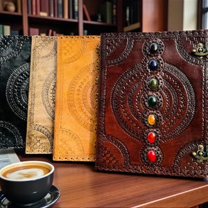 May include: A collection of leather journals in various colors, including black, tan, and brown. The brown journal features decorative stitching and colorful gemstones. A cup of coffee sits in the foreground.