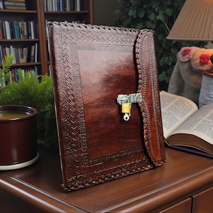 Puede incluir: Un diario de cuero marrón oscuro con un diseño floral en relieve y un candado de metal. El diario está abierto, revelando páginas en blanco. Una taza de té y un libro abierto están sobre una superficie de madera.