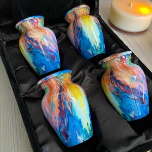 May include: Four small, colorful urns with a marbled effect, displayed in a black velvet box. The urns feature a mix of blue, yellow, red, and orange hues. A candle is visible in the background.