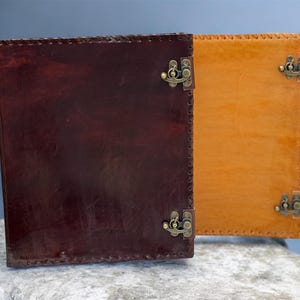 May include: Two leather journals, one dark brown and one tan, are displayed side-by-side. Each journal has a rectangular shape with metal clasp closures and stitched edges. The journals are resting on a light gray stone surface.