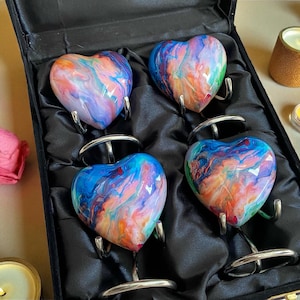 Puede incluir: Cuatro objetos decorativos en forma de corazón con patrones coloridos y arremolinados, presentados en una caja negra con forro de satén. Cada corazón está montado sobre un soporte plateado. La paleta de colores incluye azules, naranjas y morados. También se ven velas y rosas rosas.