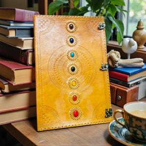 May include: A large, yellow leather journal with colorful gemstone accents and intricate embossed designs. The journal is open, revealing blank pages. A stack of vintage books and a teacup are nearby.