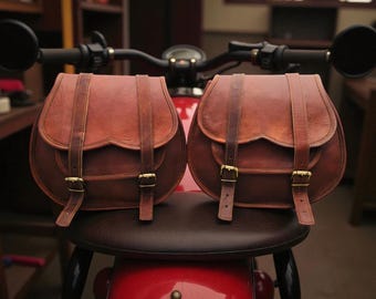May include: Two brown leather saddlebags with brass buckles and straps. The bags are mounted on a red motorcycle, with the handlebars and mirrors visible in the background. The bags have a classic, vintage design.