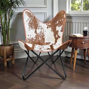 May include: Butterfly chair with a brown and white cowhide pattern seat and a black metal frame. The chair has a brown leather trim. A small wooden side table and a potted plant are in the background.