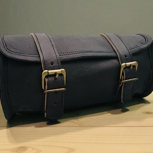May include: A cylindrical dark navy blue bag featuring two vertical straps with antique brass-toned metal buckles. The bag has a smooth, matte texture and a flap closure that secures with the adjustable buckles, creating a structured appearance.