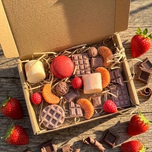 May include: A cardboard gift box filled with assorted wax melts shaped like food items, including a red macaron, chocolate bars, waffle, orange slices, and berries, nestled in paper straw filler, with fresh strawberries and chocolate pieces nearby.