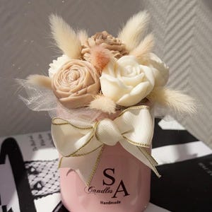 May include: A pink candle holder with a floral arrangement of cream and tan roses, and fluffy accents. A white bow with gold trim adorns the front. The text "SA Candles Handmade" is printed on the front of the candle holder.