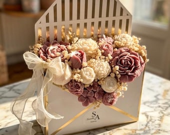 May include: A white envelope-shaped box with gold-lined stripes on the flap, filled with an arrangement of mauve and cream-colored flower-shaped candles and dried baby's breath. A sheer white ribbon is tied to the side of the envelope.
