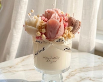 以下が含まれることがあります： Happy Birthday Margaretと書かれた白いキャンドルが入ったガラス製キャンドルホルダー。上部にはピンク、クリーム、茶色のチューリップ型のワックスフィギュアと小さなドライフラワーが飾られ、Just for youとプリントされたリボンで結ばれています。