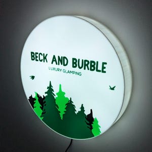 May include: A circular, illuminated sign displaying the text "BECK AND BURBLE LUXURY GLAMPING" in green. The sign features a forest silhouette and two bird graphics. A black power cord is visible at the bottom.