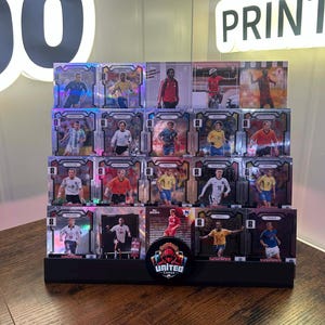 Puede incluir: Un expositor negro de varios niveles que contiene diecinueve tarjetas coleccionables de fútbol rectangulares con varios jugadores en equipaciones internacionales. Una insignia circular negra con una figura encapuchada roja y el texto UNITED CARDS está fijada en el centro frontal.