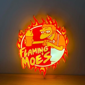 May include: A Flaming Moe's sign featuring a cartoon bartender holding a drink, surrounded by flames. The sign has a red and yellow colour scheme with the text "Flaming Moe's".