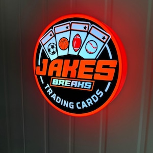 Op de afbeelding: Een rond, verlicht bord met een zwarte achtergrond en oranje rand. Het bord toont de tekst "JAKES BREAKS TRADING CARDS" en afbeeldingen van een voetbal, basketbal, American football en honkbal.