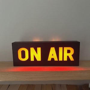 May include: A rectangular, dark brown "ON AIR" sign with bright yellow illuminated letters. The sign sits on a wooden shelf, with a soft red glow emanating from the bottom. The background is a neutral gray wall.