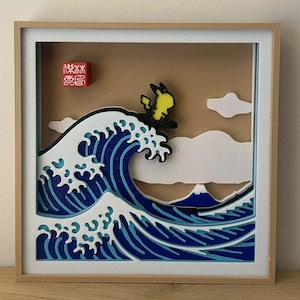 Può includere: Un'opera d'arte 3D incorniciata raffigurante un personaggio giallo che fa surf su una grande onda blu e bianca, che ricorda l'arte giapponese. Lo sfondo presenta un cielo marrone, nuvole bianche e una montagna. Un timbro rosso è nell'angolo in alto a sinistra.