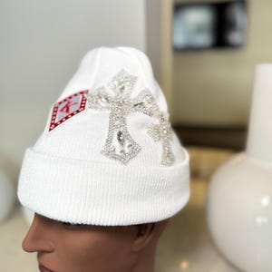 May include: White knit beanie featuring a large, sparkling cross design. The hat also has a red and white emblem on the side. The beanie is displayed on a mannequin head.